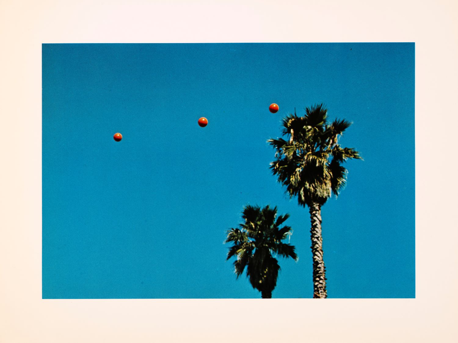 Photograph of three orange balls thrown into blue sky with palm trees.