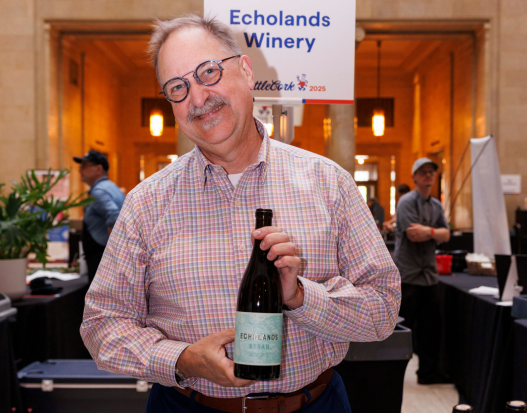 A person in glasses with a mustache holds a bottle of wine and smiles.